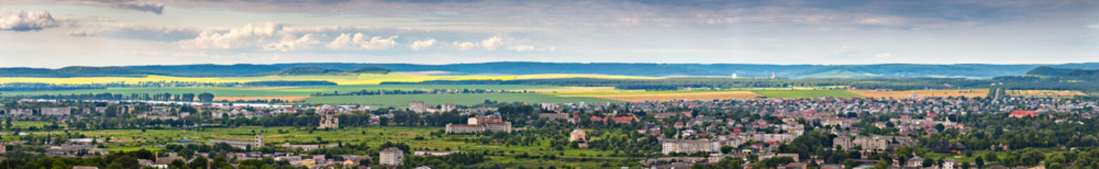 A high-angle panoramic view of the town of Zolochiv in Lviv Oblast, Ukraine. The cityscape, featuring a railway and red-roofed houses, is surrounded by green hills and agricultural fields.