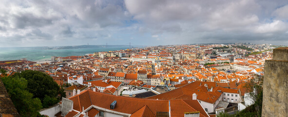 A beautiful panoramic, high-angle view of the Lisbon, Portugal cityscape. The frame includes the historic red rooftops of the old town, the wide Tagus River, the 25 de Abril Bridge under a cloudy sky.