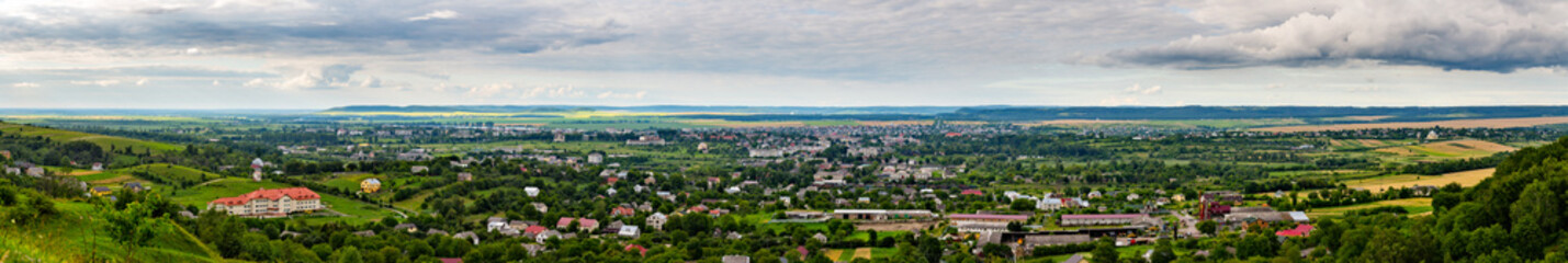 A high-angle panoramic view of the town of Zolochiv in Lviv Oblast, Ukraine. The cityscape, featuring a railway and red-roofed houses, is surrounded by green hills and agricultural fields.