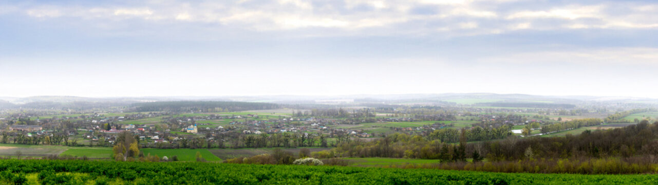 A panoramic view of Mliyiv village, Cherkasy region, Ukraine, in the Vilshanka River valley, during spring late April. The photo shows houses and buildings surrounded by green fields and rolling hills