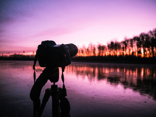 silhouette of camera standing by lake during sunset with colorful sky and calm reflections
