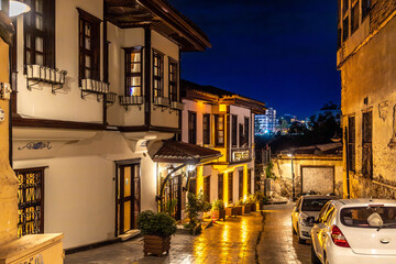Fototapeta premium A narrow street in Kaleici old town illuminated by evening lights, featuring traditional Ottoman houses with cumba balconies, flower pots, and reflections on wet cobblestones. Antalya, Turkey.