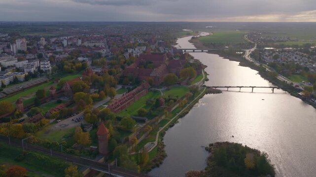 Aerial view tracks across Malbork Castle, the Nogat River, bridges, a railway, town blocks, lawns, and autumn trees under low cloudy late afternoon light.