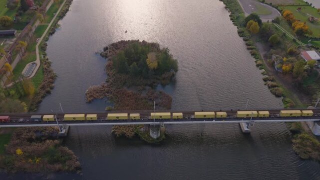 Aerial follows a long yellow and red freight train on an electrified bridge over a calm river with a marshy islet, parklike banks, and late afternoon autumn light.