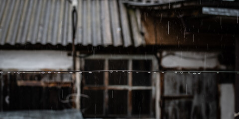 A close-up of raindrops on a clothesline against a rural backdrop. A blurred old building (barn) with a white wall and wooden window is visible.