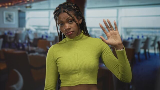 Woman with raised hand palm out in a restaurant building, cropped green top and braided hair visible; personal boundary indifference.