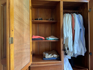 A wooden wardrobe with its door open, revealing a couple of belts on the top shelf and folded clothes on the two shelves below. A group of shirts hangs in the adjacent cubicle.