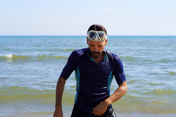 Man wearing diving goggles and a wetsuit emerges from the water after swimming in the sea.