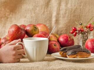 Child's hands holding a mug next to a chocolate eclair and autumn apple harvest