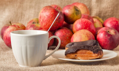 White mug and chocolate eclair in focus against a blurred backdrop of red apples.