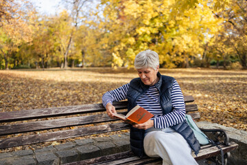 Fototapeta premium Senior woman relaxing reading book on park bench in autumn