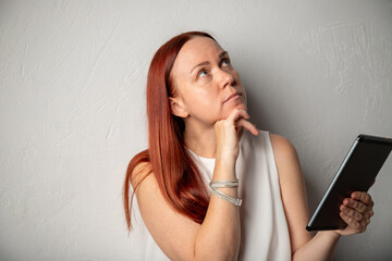 An adult Irish woman with red hair, freckles and no makeup looks up in thought, holding a tablet in her hand