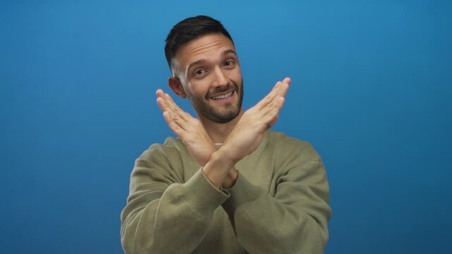 Young man in a green sweater crosses his hands in an x gesture against a solid blue background indicating rejection with a confident expression.