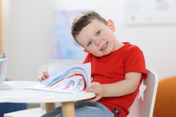 Funny little boy with child's drawing of family and rainbow at white table indoors