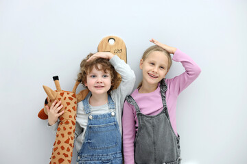 Little boy and girl with toy giraffe measuring their height with chart near light grey wall
