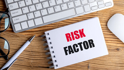 Notebook with text risk factor on wooden desk with keyboard, pen, glasses and computer mouse