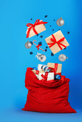 Christmas balls, gift boxes and candy cane falling into bag on blue background