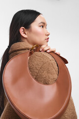Asian woman wearing a warm brown coat shows a side profile as she carries a round leather bag over...