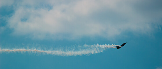 A fighter plane is flying in the cloudy sky with a trail of smoke that curls behind it. The wide perspective impressively reflects the elegance of flight and the vastness of the sky.