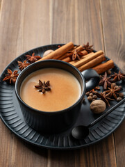 Traditional Indian masala chai tea with milk and spices in a black cup on a dark plate on a wooden background