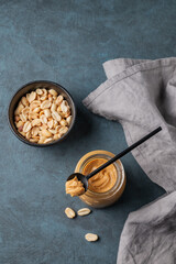 Creamy peanut butter in a jar on a dark background with roasted nuts spoon and napkin.