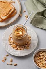 Creamy peanut butter in a jar on a light background with toasted bread, scattered roasted nuts
