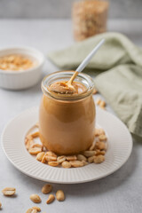 Creamy peanut butter in a jar on a light background with scattered roasted nuts, spoon and napkin