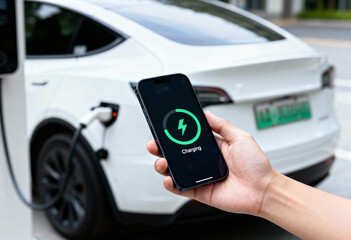 Hand holding phone displaying charging status, near white electric vehicle connected to charging station.