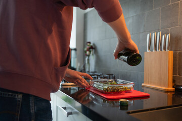 Person in casual attire pouring olive oil over fresh vegetables in a glass dish on a modern kitchen countertop, showcasing healthy cooking and meal preparation techniques