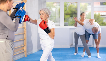 Active aged lady wearing boxing gloves practicing punches with trainer during sports classes