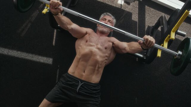 Top-down view of senior Caucasian man with muscular body doing bench press exercise with barbell in gym