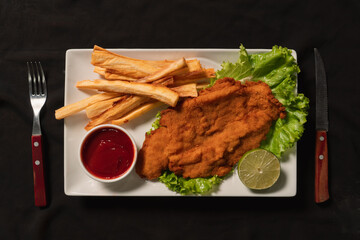 Top view of breaded cutlet with French fries and ketchup served on a white plate with cutlery.