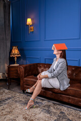 Fototapeta premium Woman covering her face with a book while resting on a luxurious leather sofa, depicting study break or mental exhaustion