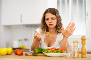 Young sad grimacing and gesturing woman eating healthy vegetable salad at kitchen, eating disorder