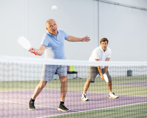 Portrait of concentrated experienced aged pickleball player preparing to strike and return ball to opponents field during doubles match in team with male partner on indoor court