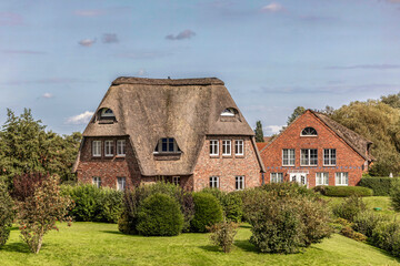 Typical North German architecture near Glückstadt with historic red brick houses and thatched roofs, surrounded by lush green garden and trees under a clear summer sky in Schleswig-Holstein.