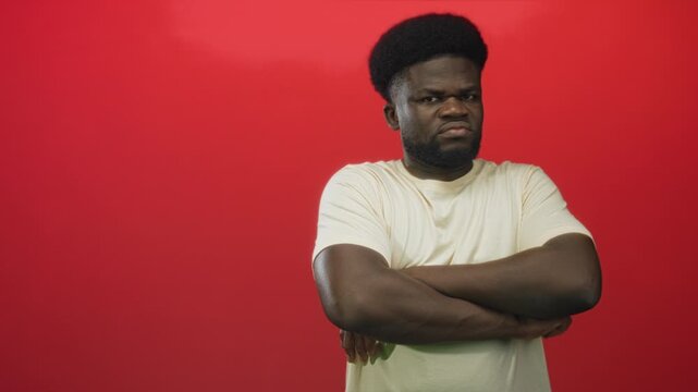 Young african american man in beige t shirt standing with arms crossed and a frown against a bold red studio wall; defiance.