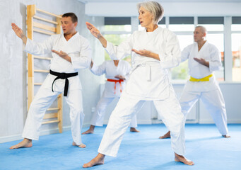 Diligent old man attendee of karate classes practicing kata standing in row with others in sports hall