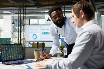 Black male manager with caucasian coworker planning new business initiatives, reviewing market analytics. Businessmen analyzing stock trading performance to strengthen achieve organizational success.