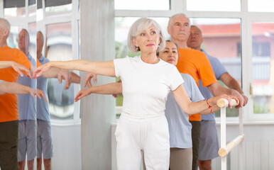 Group of positive aged women and men standing in row together while doing ballet dance moves, dancers standing in 2nd position of ballet stance in bright fitness room