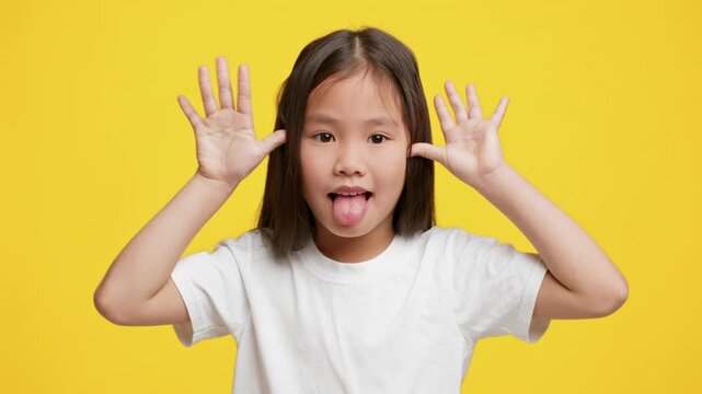 A playful Asian child expresses joy by sticking out her tongue and making silly gestures. The vibrant yellow background amplifies the sense of fun and carefree childhood moments.