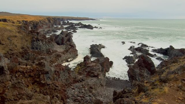 An aerial or wide angle shot of the rugged coastline of Iceland's Sn&aelig;fellsnes Peninsula. The scene features dramatic volcanic rock formations and cliffs against a turbulent sea with white foam from cr