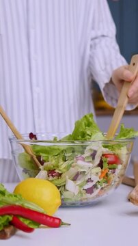 Woman in pajamas adding fresh lettuce into a glass bowl and mixing a healthy vegetarian salad with wooden spoons on a kitchen table with vegetables, bread, and juice in the background