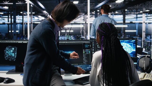 Multiracial technicians in diverse data center review AI diagnostics, analyzing neural network performance. Asian man and african american woman optimize artificial intelligence systems, camera A