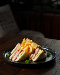Club Sandwich with Ham, Egg, and Vegetables Served with Potato Chips on Black Plate
