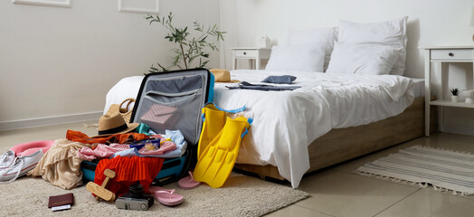 Open suitcase with clothes and beach accessories on carpet in bedroom interior