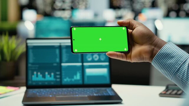 Black professional looks at chroma key on mobile phone screen at desk, solving business management tasks after hours. Male staff oversees project development, company objectives. Camera B.