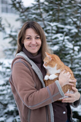 Woman holds a ginger cat in a basket while surrounded by snow-covered trees during winter celebrations