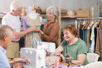 Enthusiastic aged woman stitching at machine in tailoring studio, enjoying hands-on learning during sewing class for older adults, while fellow learners sharing creative work and friendly conversation