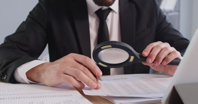 Young man auditor checking financial documents with magnifying glass over office desk. Fraud investigation and tax audit. Male accountant searching information in document. 4k video.